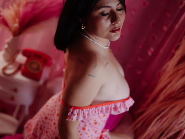 Sara Guzman, met een gemiddelde borstmaat, draagt een rode en witte jurk met stippen, een off-shoulder model, een parelketting en een korte zwarte haar, leunt voorover, en toont tatoeages op haar rechter schouder, in een roze kamer.