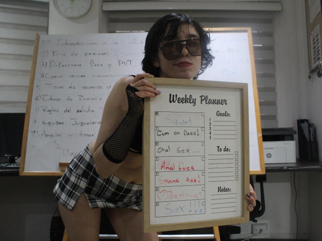 Elodie Perrine staat in een donker kantoor, een wekelijkse agenda in haar handen. Ze draagt een zwart top met stippen, een geruite rok, een bril en heeft een rondingen. De achtergrond toont een whiteboard met handgeschreven tekst..