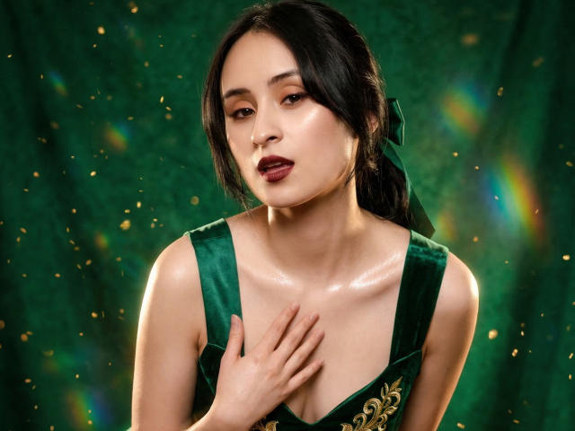 Annie Lamberth, une femme asiatique attrayante avec de longs cheveux noirs, porte une robe en velours vert avec des broderies dorées, qui la recouvre. Elle posa une main sur sa poitrine. L'arrière-plan est vert avec des taches de lumière colorées. Ses lèvres sont d'un rouge foncé, son expression est séduisante..