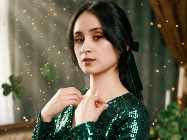 Annie Lamberth, una donna asiatica con la pelle chiara e i capelli neri, guarda seducentemente l'obiettivo. Indossa un abito verde scintillante ricoperto di paillettes, con le mani vicine al petto. La scintillante luce la circonda.
.