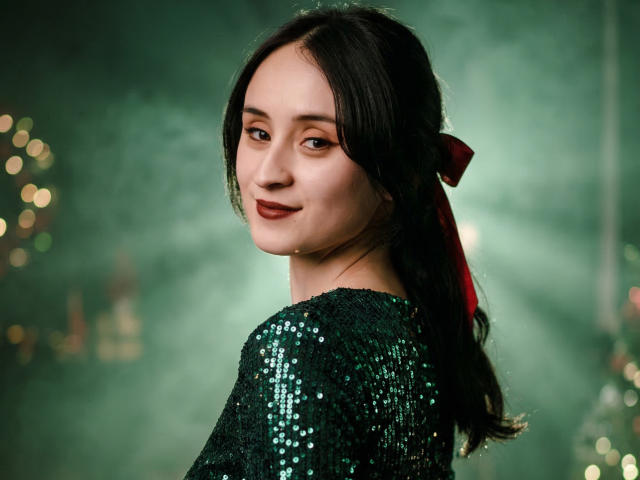 Annie Lamberth, de costas, mas olhando por cima do ombro, vestindo um vestido preto com lantejoulas e um laço vermelho em seu cabelo escuro. cabelo, com uma luz verde brilhante atrás dela, sorriso suave, ar sensual..