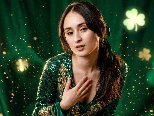 AnnieLamberth with long dark hair, wearing a green sequined top, hand on chest, green and gold pattern, glowing yellow particles in dark background, intense gaze, sexual tension, close-up portrait.