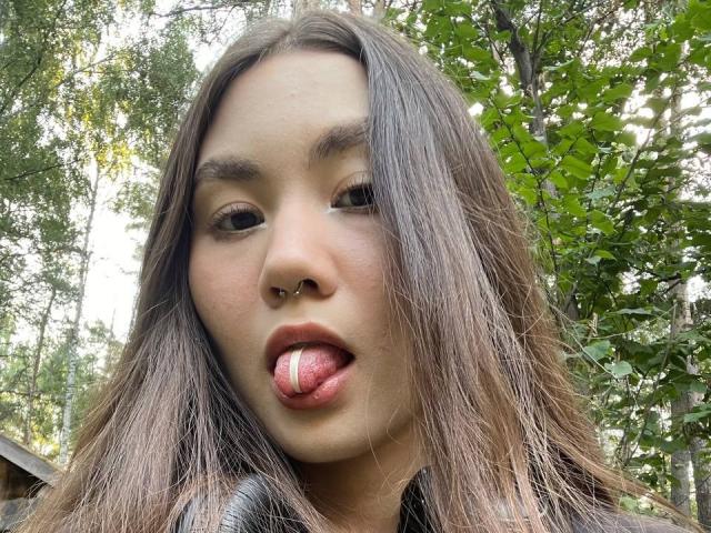 Yolande Asmar heeft lang bruin haar, een neusring en steekt haar tong uit met een roze en witte lolly erop. Ze is buiten, met groene bomen op de achtergrond..