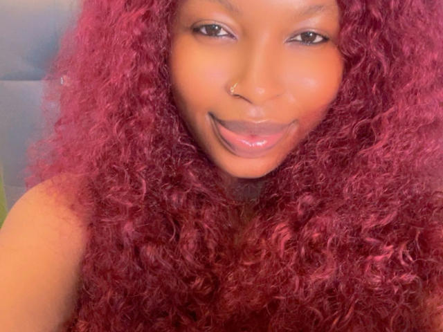 Selfie de primer plano de Annykaa con cabello rizado rojo vibrante, sonriendo con labios brillantes, un aro en la nariz y piel oscura. Ojos brillantes, una ligera sombra en la mejilla, una vibra íntima y sexy..