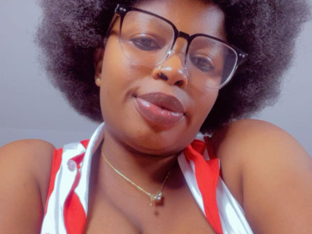 Gros plan d'Annykaa, avec une peau foncée, une grande coiffure afro et des lunettes à monture épaisse, portant un haut à encolure américaine rouge et blanc. Elle regarde la caméra, les lèvres légèrement entrouvertes, avec un sourire discret..