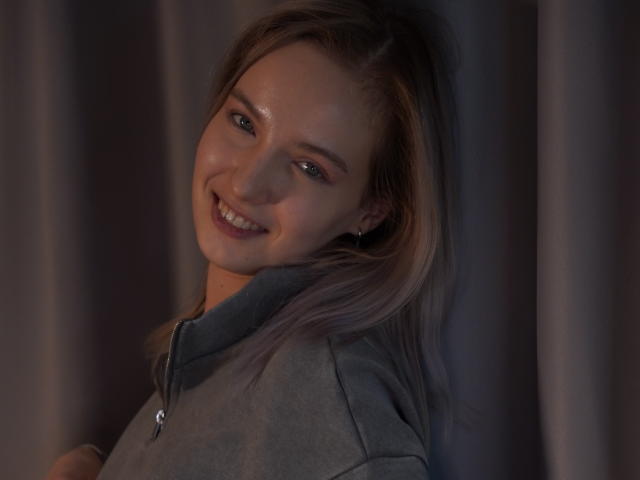 Fotografia de Nicole Spritzer com cabelo castanho claro, sorrindo com um leve inclinação da cabeça, vestindo uma jaqueta cinza, com pouca luz. fundo escuro, suor visível no rosto dela, tensão sexual.