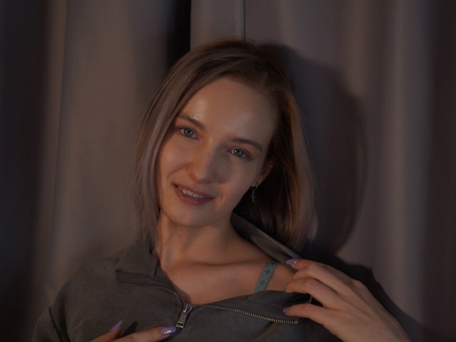 Foto van Nicole Spritzer, een lichtgetinte vrouw met bruin haar dat tot op haar schouders reikt, die verleidelijk glimlacht en een gedeeltelijk opgeritst, donkergrijs topje, waardoor een stukje blauwe bh zichtbaar is. Zacht licht, schaduwen die haar gezicht accentueren.