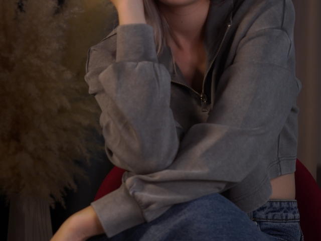 Fotografía de primer plano de NicoleSpritzer con una sudadera gris, con la mano apoyada en su rostro, y pantalones vaqueros azules. Está sentada mostrando su escote, creando un ambiente sexy y casual. Iluminación suave.