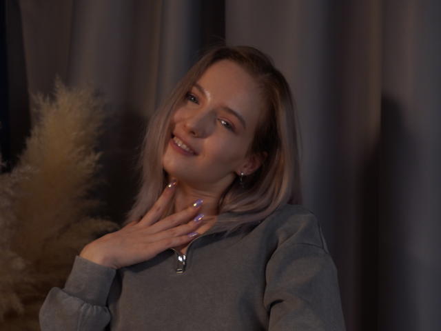 Fotografia de Nicole Spritzer, loira, sorrindo, com a mão no pescoço, usando um moletom cinza, com unhas roxas, com pouca luz, sombras suaves. Cortinas ao fundo, luz quente à esquerda, pose casual, tensão sexual sutil..