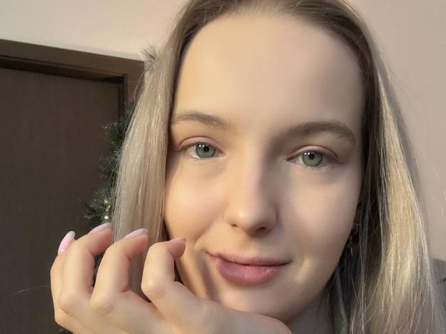 Primer plano de Nicole Spritzer, de piel clara, con ojos verdes, cabello rubio y una ligera sonrisa. Ella está tocándose la mejilla con su mano derecha, mostrando uñas largas y bien cuidadas. Expresión muy tierna. Iluminación suave..