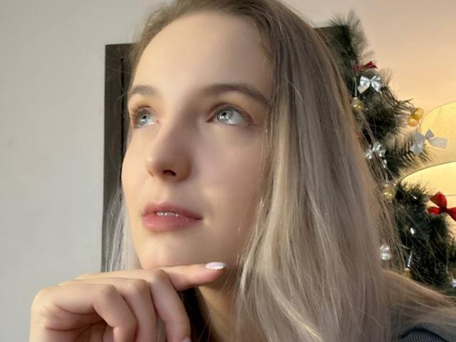 Nahaufnahme von NicoleSpritzer, die mit einem leichten Lächeln nach oben schaut, eine Hand berührt ihr Kinn, langes blondes Haar, blaue Augen. weiches Licht, Weihnachtsbaum mit Dekorationen im Hintergrund, sexy, verführerische Atmosphäre.