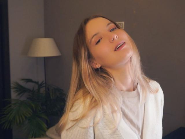 Foto van Nicole Spritzer met lichtbruin haar, leunend achterover met een verleidelijke uitdrukking, gekleed in een witte blouse. Zacht licht, lamp op de achtergrond, groene plant. Seksuele spanning, ogen half gesloten, mond licht open..