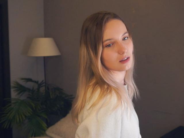 Fotografie von Nicole Spritzer mit hellbraunen Haaren, die über ihre Schulter schaut und verführerisch lächelt. Sie trägt ein weißes Oberteil, das Licht einer Lampe beleuchtet ihr Gesicht, der Hintergrund ist ein dunkler Raum. Verdammt heiß..