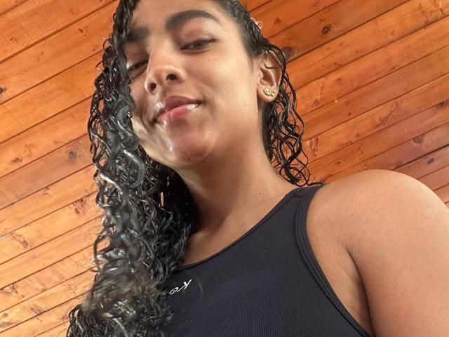 Close-up of NatalyBlonder, wet curly black hair, black tank top, wooden wall background. Her face and neck have visible semen stains. She's smiling slightly, looking at the camera. Casual, erotic vibe.