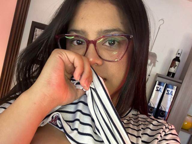 Close-up photo of ConnySaenz, holding a striped shirt to their mouth with a hand, wearing red glasses, black hair, light skin, casual striped shirt, bottle and shelves in background. Sexual tension.