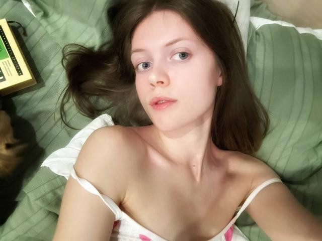 Photograph of HanHeffren lying on a green-striped bed, looking up with pale skin, blue eyes, and dark brown hair. White off-shoulder top, soft light, relaxed expression, cat partially visible.