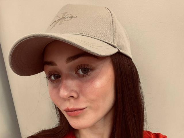 Nahaufnahme von JoanAlkinss, die eine weiße Baseballkappe trägt, mit langen braunen Haaren, braunen Augen und einem leichten Lächeln. Ihre Haut ist hell, und sie trägt ein rotes Hemd. Verdammt süß..