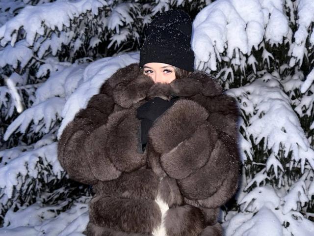 JoanAlkinss, gekleidet in einen dicken, dunkelbraunen Pelzmantel und eine schwarze Mütze, steht inmitten von schneebedeckten Kiefern. Ihr Gesicht ist teilweise sichtbar, mit Schneeflocken auf ihren Wangen. Verdammt kalt..