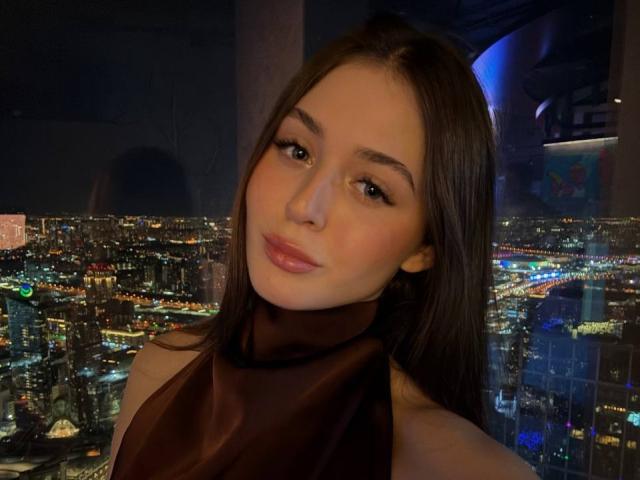 Joan Alkinss, mit langem, dunklem Haar, vollen Lippen und einem verführerischen Blick, trägt einen schwarzen Rollkragenpullover und steht in einem Hochhaus. Nacht.
die Stadtlichter beleuchten den Hintergrund und erzeugen eine sinnliche, urbane Atmosphäre..