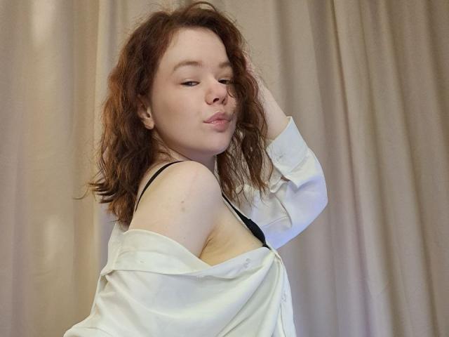 Photograph of CoreyRounsaville with messy brown hair, white shirt slipping off shoulder, black bra strap visible, looking seductively at camera with hand in hair. Background is beige curtains.