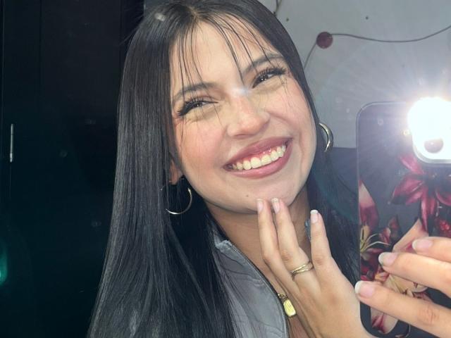 Photo en gros plan de NyraVanel, avec de longs cheveux noirs, souriant largement, montrant ses dents, tenant un téléphone avec une coque à motifs floraux. Elle portait des boucles d'oreilles pendantes et une bague en or à son doigt. Éclat lumineux.