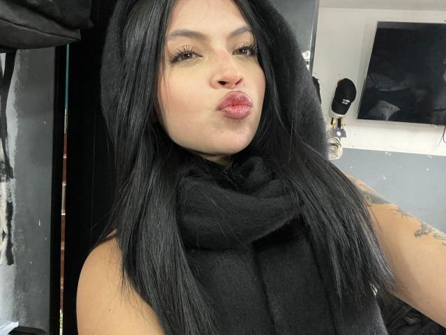 NyraVanel, a curvy woman with long black hair, puckering her lips in a kiss. She's wearing a black scarf, exposing a tattoo on her upper arm. She's giving a seductive look.
