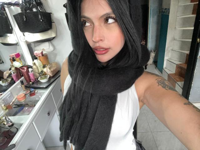 NyraVanel, con cabello negro, vestida con una camiseta blanca y un pañuelo negro, mirando por encima del hombro, revelando un tatuaje en su brazo superior. Mesa de maquillaje con productos en el fondo.