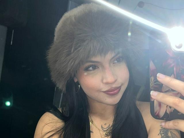 Close-up selfie of NyraVanel, wearing a gray fur hat, black hair, dark lipstick, smiling, holding a phone with a floral case, visible tattoo on right arm, bright flash on the right.
