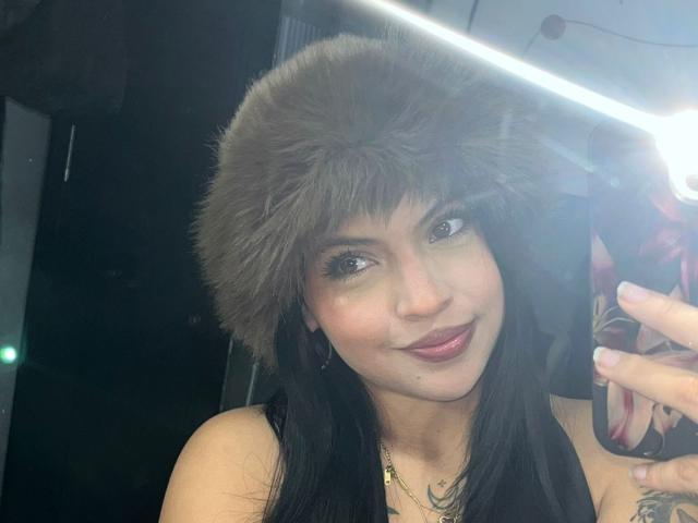Close-up photo of NyraVanel, a sexy woman with long black hair, wearing a gray fur hat, taking a selfie with a glowing flash. She has dark eyeliner, pink lipstick, and a tattoo on her right shoulder. Her expression is confident and slightly seductive.