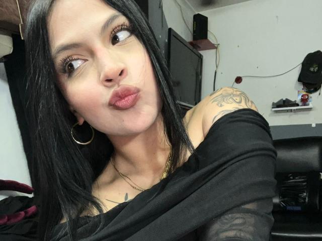 Close-up of NyraVanel, a Latina woman with long black hair, pouty lips, and tattooed shoulder, wearing a black off-shoulder top, looking seductively to the side. Background includes a chair and shelf.