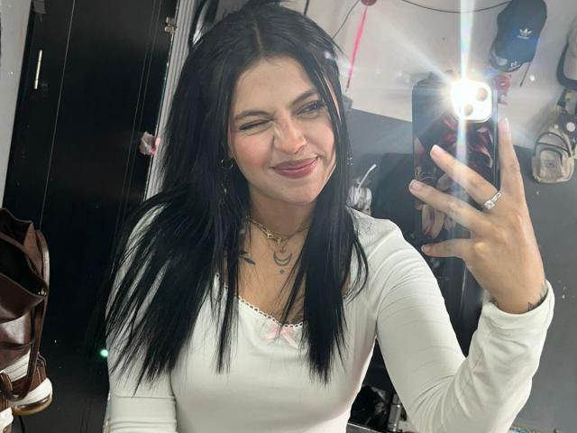 NyraVanel prend un selfie devant un miroir, avec un flash lumineux, en souriant et les yeux fermés. Elle a de longs cheveux noirs, un haut blanc et un collier. Sa main tient un téléphone, qui affiche une bague..