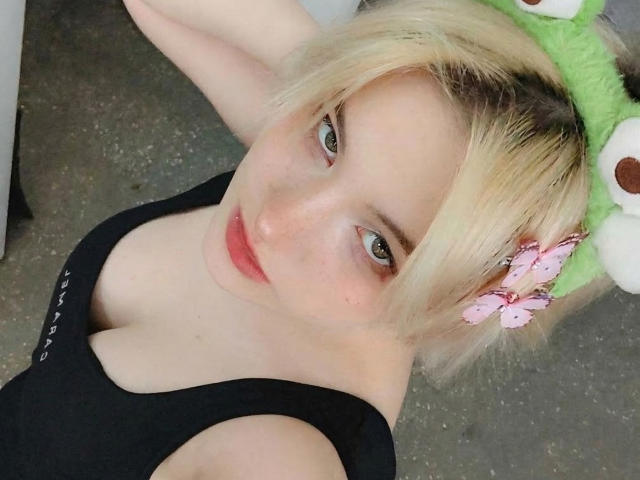 Foto ravvicinata di KatlynRenteria, bionda con un cappello a forma di rana verde, un fiore rosa, un top nero, scollatura, sguardo seducente. pelle chiara, labbra rosse, braccia alzate, sfondo grigio, aspetto casual, atmosfera sensuale..