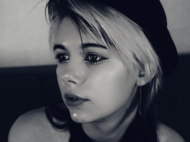Close-up photograph of LaurenceCloud with short, messy blonde hair, wearing a black cap, gazing downward with glossy, wet skin, visible droplets on face, intense blue-black monochrome filter.