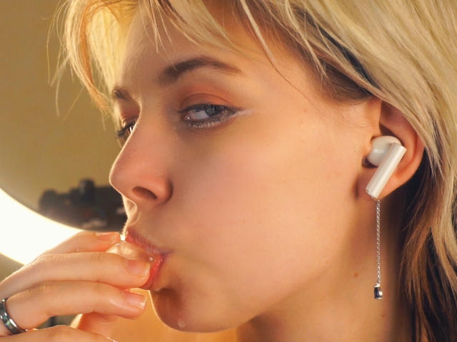 Primer plano de LaurenceCloud con cabello rubio, chupándose el dedo, usando audífonos blancos con una cadena colgante. La luz resalta su piel clara, y llevan un anillo en el dedo. Increíblemente atractivo/a..