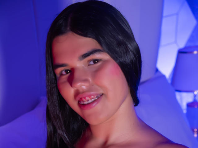 Gros plan de MiluJones, une femme séduisante avec de longs cheveux noirs, un blush rose sur les joues et un léger sourire, éclairée par une lumière bleue. éclairage. Elle est nue sur le buste, laissant apparaître des épaules lisses et une clavicule..