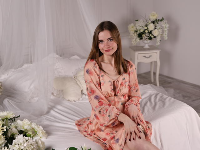 Margarita Kis, eine hellhäutige Frau mit glattem, braunem Haar, sitzt auf einem weißen Bett und trägt ein blumiges, pfirsichfarbenes Kleid. Sie hat ein leichtes Lächeln, im Hintergrund sind weiße Blumen und eine Vase zu sehen. Verdammt heiß..