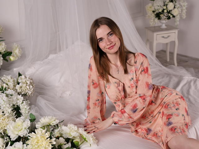 MargaritaKis, con i capelli castani e la pelle chiara, è seduta su un letto bianco, indossa un abito floreale color pesca e guarda verso la fotocamera. Con un leggero sorriso, circondato da fiori bianchi. Tensione sessuale.