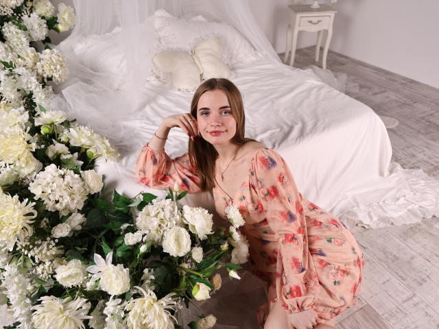 Margarita Kis arrodillada junto a una cama, vestida con un vestido rosa con estampado floral, rodeada de flores blancas, con una expresión sensual y seductora. expresión. La luz del sol ilumina su rostro y la ropa de cama blanca que hay detrás..