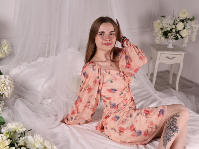 MargaritaKis, een lichtgetinte vrouw met bruin haar, gekleed in een bloemenjurk in perzikkleur, zittend op witte satijn lakens, met haar hand op haar hoofd, tatoeage op de dij, bloemen op de voorgrond, witte bijzettafel met vaas op de achtergrond. Heel aantrekkelijk..