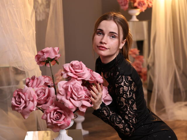 MargaritaKis with light brown hair, wearing a black lace dress, holding pink roses in foreground. Soft light, blurred background with beige curtains. Slight smile, focused gaze, sexy pose.