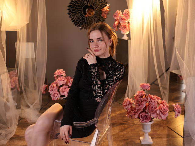 MargaritaKis, una donna dalla pelle chiara e con i capelli castani, è seduta su una sedia trasparente, indossando un abito nero in pizzo, con le gambe incrociate. Mano sul mento.
rose rosa, lampade accese, tende leggere, parete scura. Incredibilmente sexy..