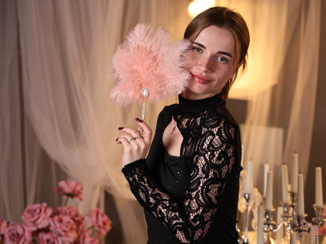 MargaritaKis, com cabelo castanho claro, vestindo uma blusa preta de renda, segurando um espanador de penas rosa em frente ao rosto, sorrindo. O fundo tem velas e rosas cor-de-rosa. Há tensão sexual devido à pose provocante..