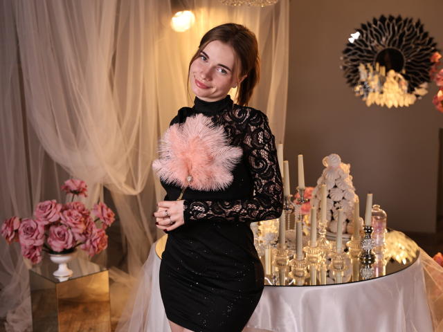 MargaritaKis se tient devant une table sur laquelle sont posées des bougies allumées et des roses roses, vêtue d'une robe noire en dentelle avec des détails roses. Détail de la poitrine ornée de plumes. Elle sourit, avec un regard coquin..