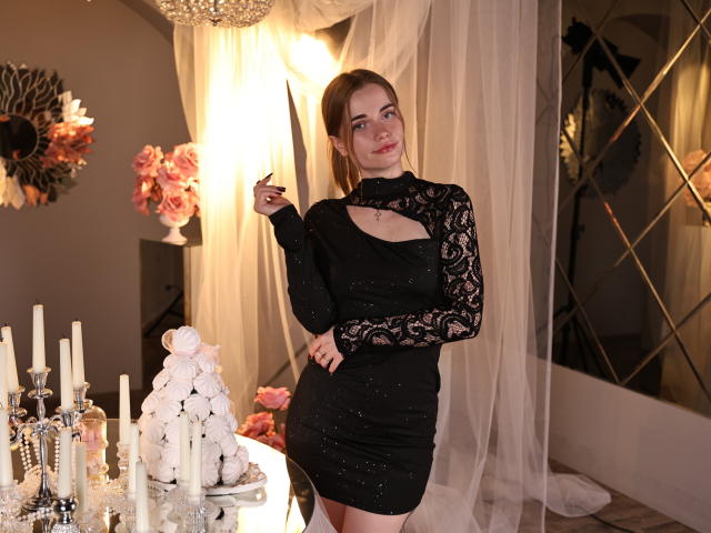 Margarita Kis está de pie en una habitación con poca luz, vestida con un vestido ajustado de encaje negro con mangas largas, que deja ver parte de su escote. Ella sostiene una vela encendida, rodeada de velas blancas. Pose seductora..