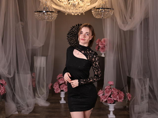 MargaritaKis è in piedi con sicurezza, indossando un abito nero a maniche lunghe con dettagli cut-out, circondata da rose rosa e cristalli. lampadari. Ha uno sguardo seducente, con una mano appoggiata sul fianco, che trasmette una forte sensualità..