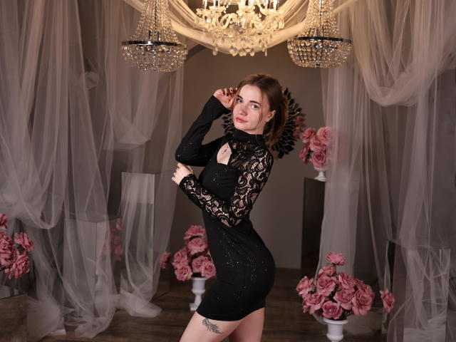 Margarita Kis, vestida con un ajustado vestido negro de encaje, está de pie en una habitación tenuemente iluminada, con un candelabro de cristal sobre ella. Ella tiene un tatuaje en el muslo, rodeado de rosas rosadas. Increíblemente sexy..