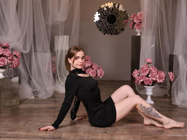 MargaritaKis está sentada no chão de madeira, com as pernas abertas, vestindo um vestido preto, que revela uma tatuagem na coxa direita. Rosas cor-de-rosa em vasos brancos, espelho com design de raios solares pretos ao fundo. Pose incrivelmente sexy..