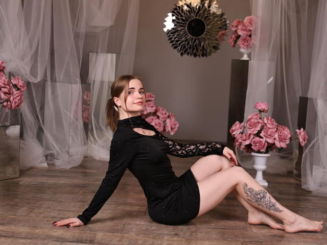 MargaritaKis está sentada em um chão de madeira, com as pernas cruzadas, vestindo um vestido preto de manga comprida com detalhes em renda. Ela tem uma tatuagem na coxa direita. Rosas cor-de-rosa e um sol negro em forma de explosão são refletidos ao fundo. Sua expressão é confiante, com um leve sorriso. Seu braço direito está atrás dela, a mão esquerda no chão..