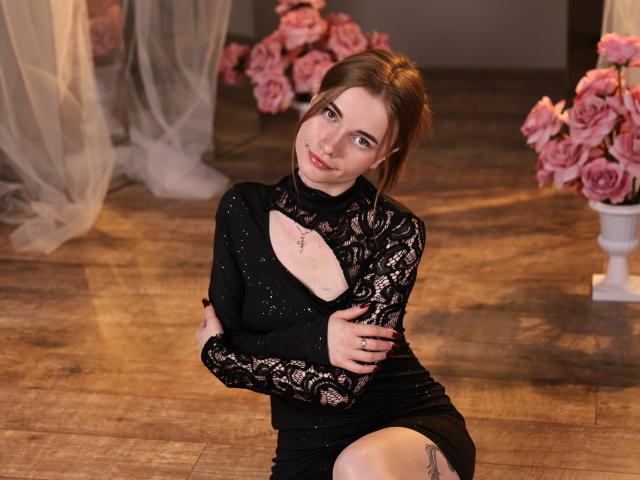 Margarita Kis, vestida con un vestido negro de encaje con un corte, está sentada en un suelo de madera, abrazándose el brazo, con una sonrisa seductora. rodeado de rosas rosadas en el fondo.