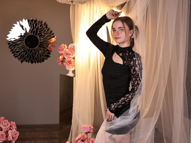 MargaritaKis è in piedi in una stanza scarsamente illuminata, indossa una maglia nera in pizzo a maniche lunghe, con tende trasparenti drappeggiate intorno a lei. Lei alza un braccio, mettendo in mostra la sua figura slanciata. Sullo sfondo, ci sono rose rosa e uno specchio nero a forma di sole..